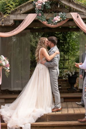 Romantic wedding ceremony kiss under wooden arbor decorated with blush drapery and flowers.