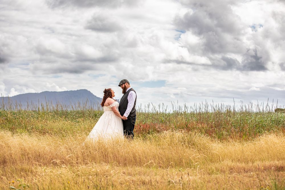Bride and groom stand facing each other in golden field with mountain backdrop and dramatic cloudy sky.