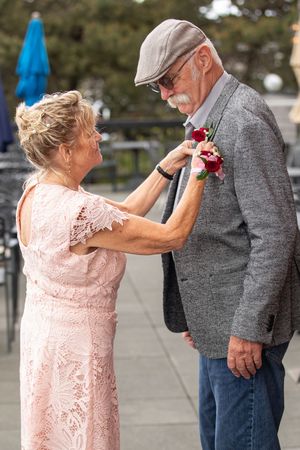 Elderly woman in pink lace dress pinning a red rose boutonniere on a man in a gray blazer and flat cap.