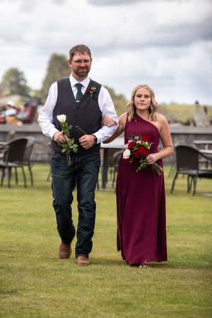 A man in a vest and woman in a burgundy gown walk arm-in-arm holding roses at an outdoor wedding ceremony.