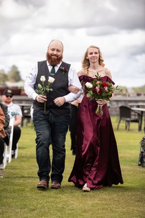 A bearded groomsman in a vest walks a bridesmaid in a burgundy gown down the aisle at an outdoor wedding.