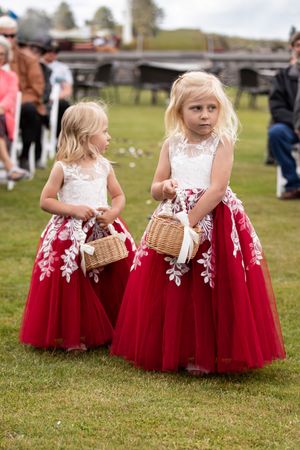 Two blonde flower girls in red and white dresses carrying wicker baskets at an outdoor wedding ceremony.