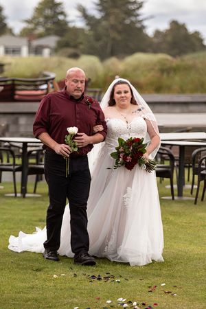 Bride in white lace gown with bouquet walks down aisle with bald man in burgundy shirt at outdoor wedding.