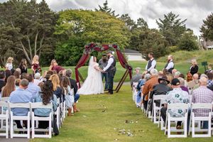 Outdoor wedding ceremony on green lawn with couple at floral arch, guests seated in white chairs under cloudy sky.