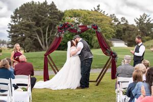 Bride and groom share first kiss under burgundy hexagonal arch with floral decor at outdoor wedding ceremony on green lawn.