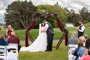 Bride and groom sharing first kiss under burgundy floral arch at outdoor wedding ceremony on lush green golf course.