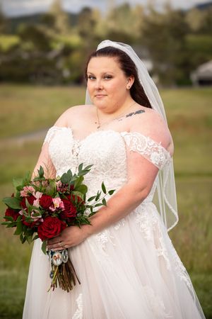 Bride in off-shoulder lace wedding gown and veil holds red rose bouquet outdoors in green field.