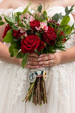 Bride holding a lush red rose and pink floral wedding bouquet with greenery, adorned with memorial photo charms.