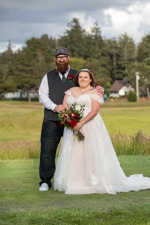 Bride in white lace gown holding red bouquet poses with bearded groom in vest on green lawn at outdoor wedding.