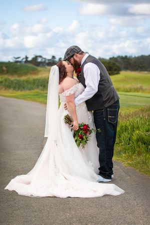 Bride and groom sharing a romantic kiss on a path, bride in white gown with veil, groom in vest and jeans.