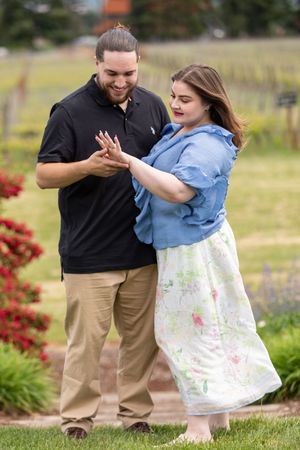 Couple admiring an engagement ring outdoors at a vineyard, woman in blue top and floral skirt, man in black polo.