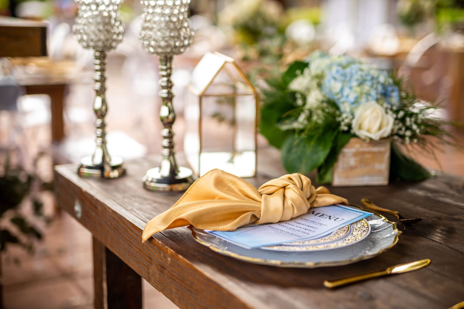 Elegant wedding table setting with crystal stemware, gold napkin and blue floral centerpiece.