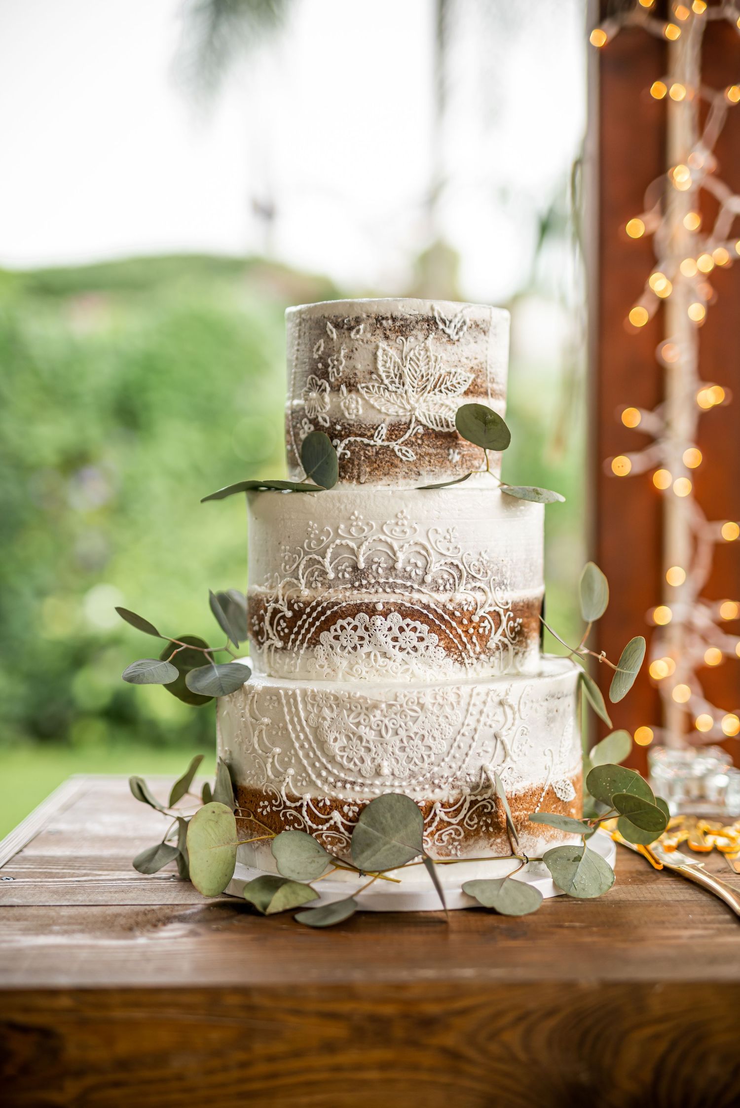 Three-tiered white wedding cake decorated with lace patterns and eucalyptus leaves.