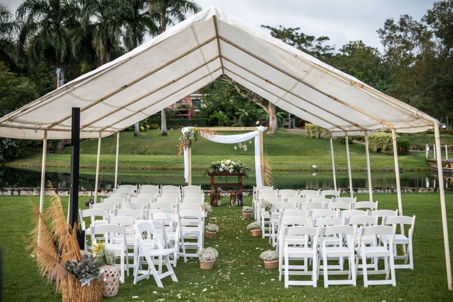 An outdoor wedding ceremony setup with white chairs and a tent canopy on a grassy lawn surrounded by palm trees.