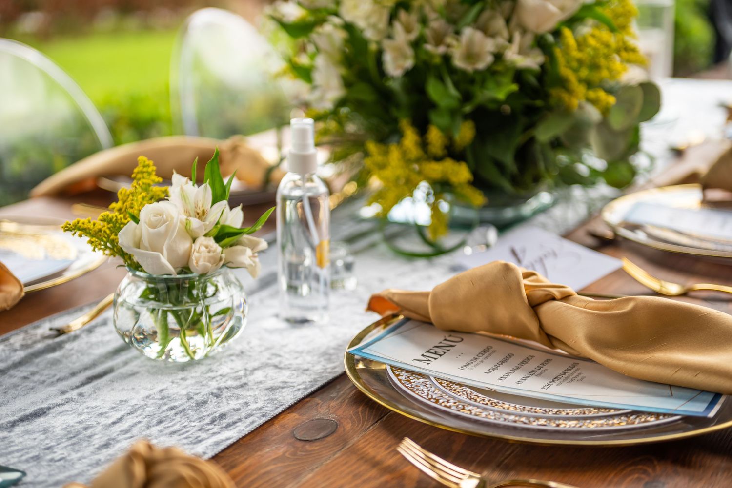 Elegant wedding table setting with floral centerpiece, gold napkins and crystal glassware on lace tablecloth.