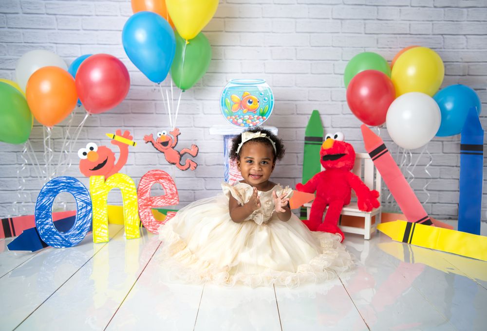 Baby girl in a colorful Sesame Street themed children's party setup with balloons, decorations and plush Elmo against white brick wall.