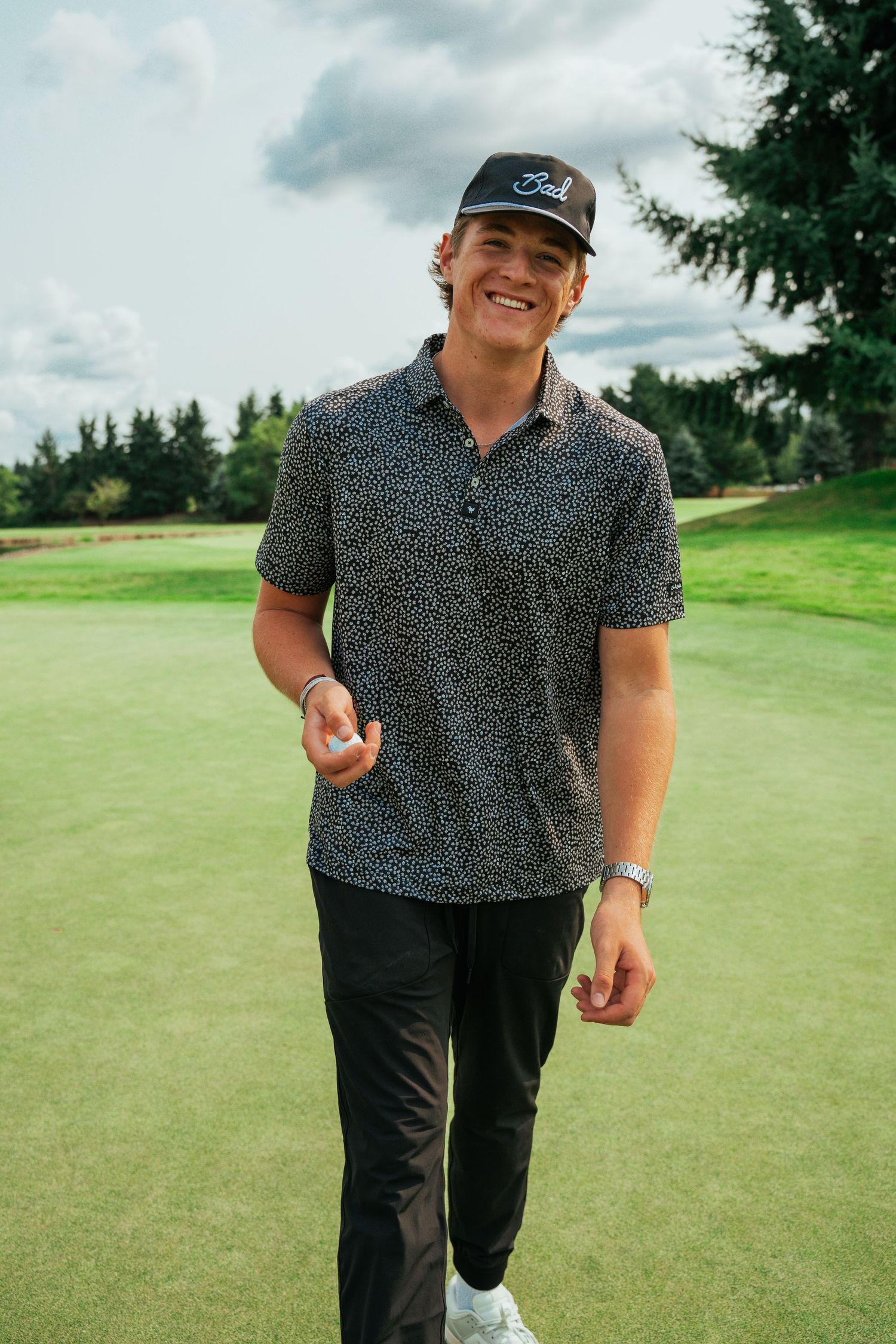 Golfer standing on green course wearing patterned polo and baseball cap.