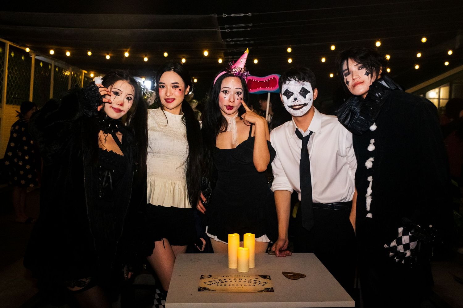 A group in Halloween costumes including dark outfits and face paint pose together at a nighttime party with string lights above.