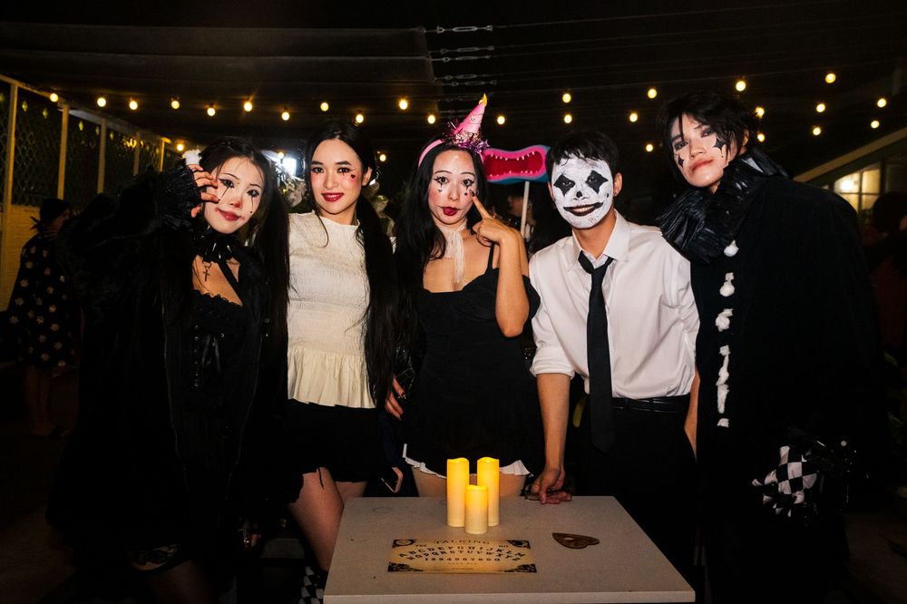 A group in Halloween costumes including dark outfits and face paint pose together at a nighttime party with string lights above.