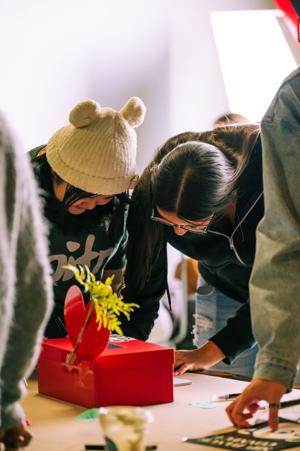 People gathered around a table with a red gift box and yellow flower, leaning in to write or examine something.