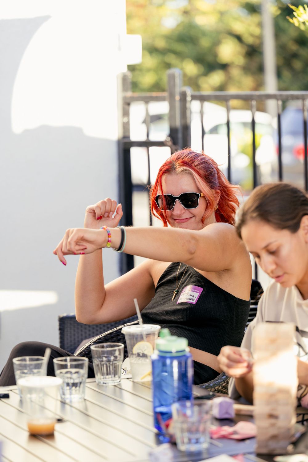 DJ with vibrant red hair and sunglasses enjoying a sunny outdoor patio with drinks on the table.