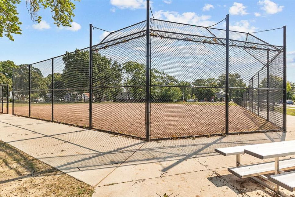 Chain-link fenced baseball diamond with dirt infield and bleachers in a sunny park setting.