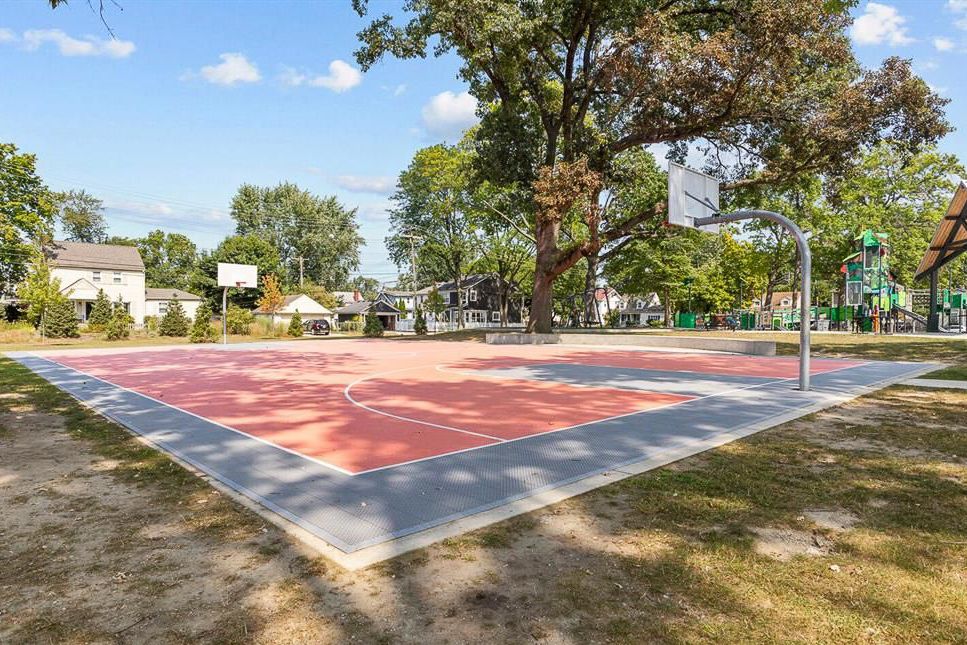 Outdoor basketball court with red and gray surface surrounded by mature trees in a public park.