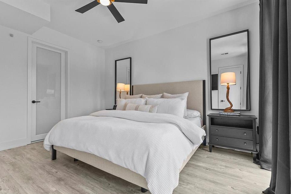 Bright modern bedroom with king bed, ceiling fan, mirrored nightstands, and light hardwood floors.