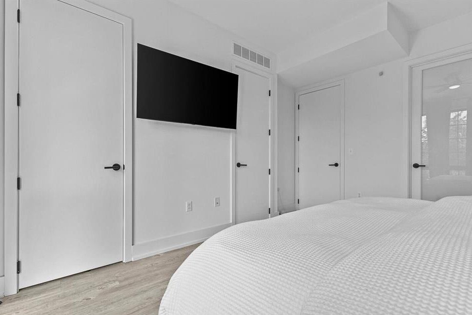 Minimalist white bedroom with wall-mounted TV, multiple closet doors, and light wood flooring.