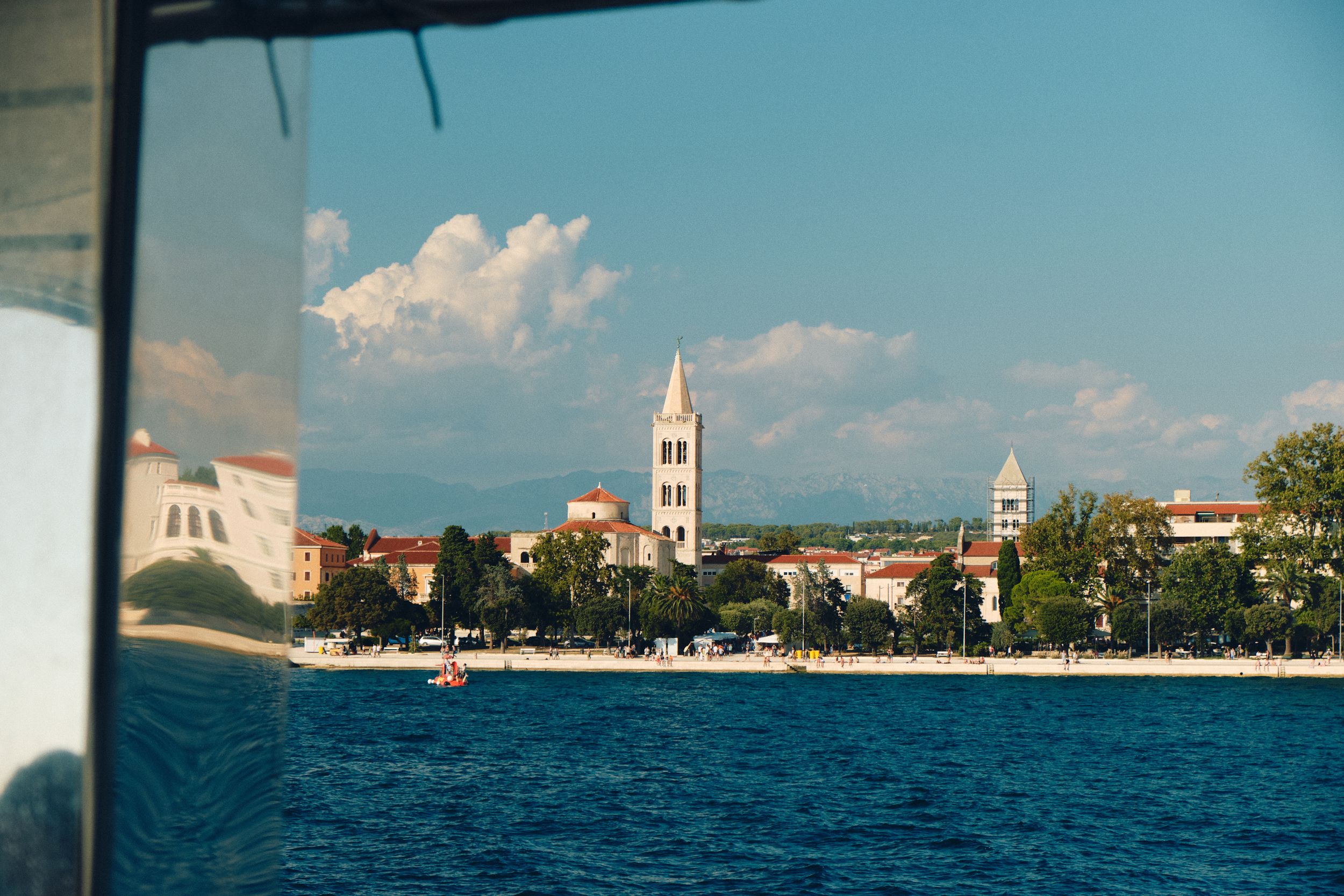 Coastal town skyline with church towers and buildings along a sunny Mediterranean waterfront.