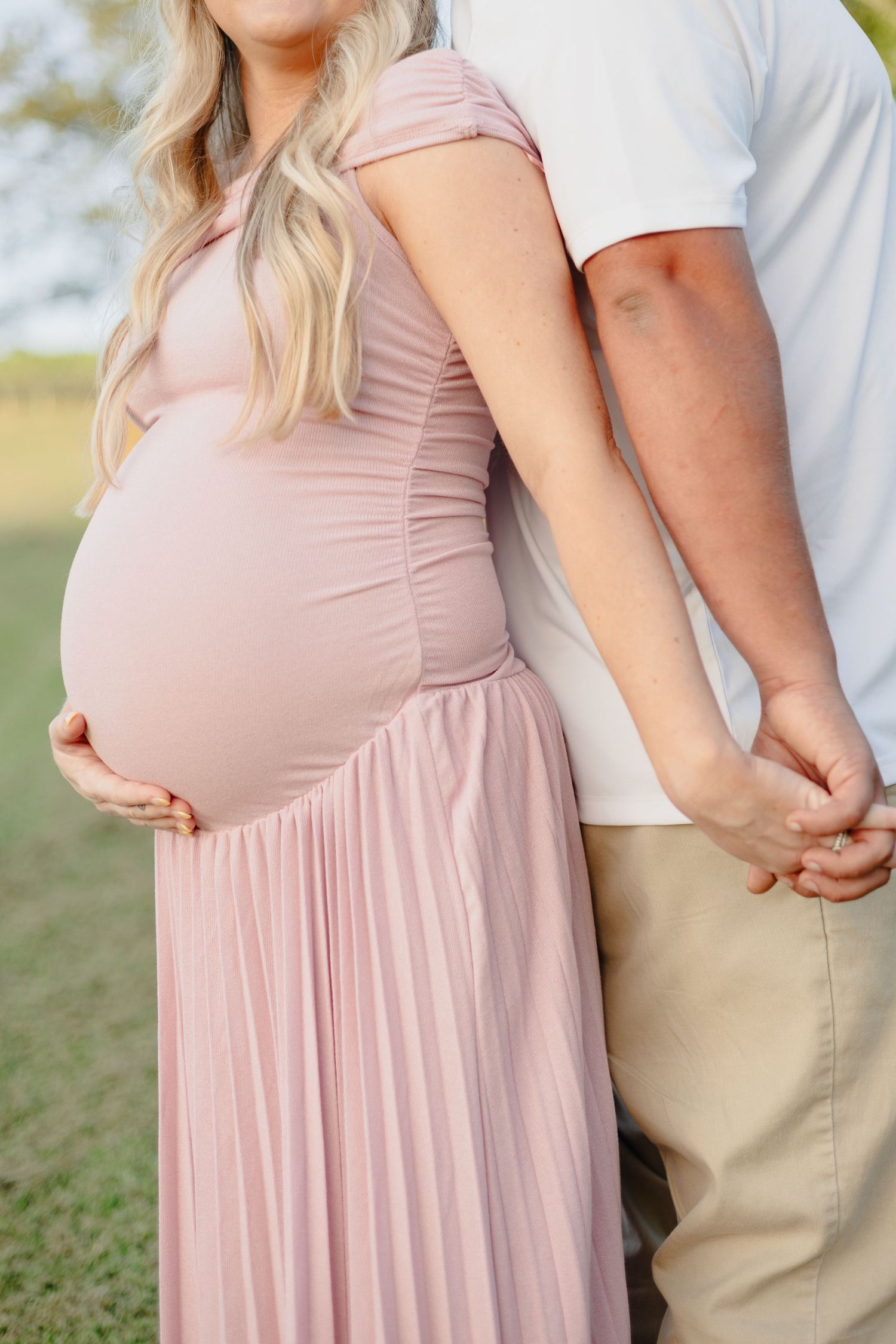 An expectant mother in a flowing off-shoulder dress stands in a sunlit garden.