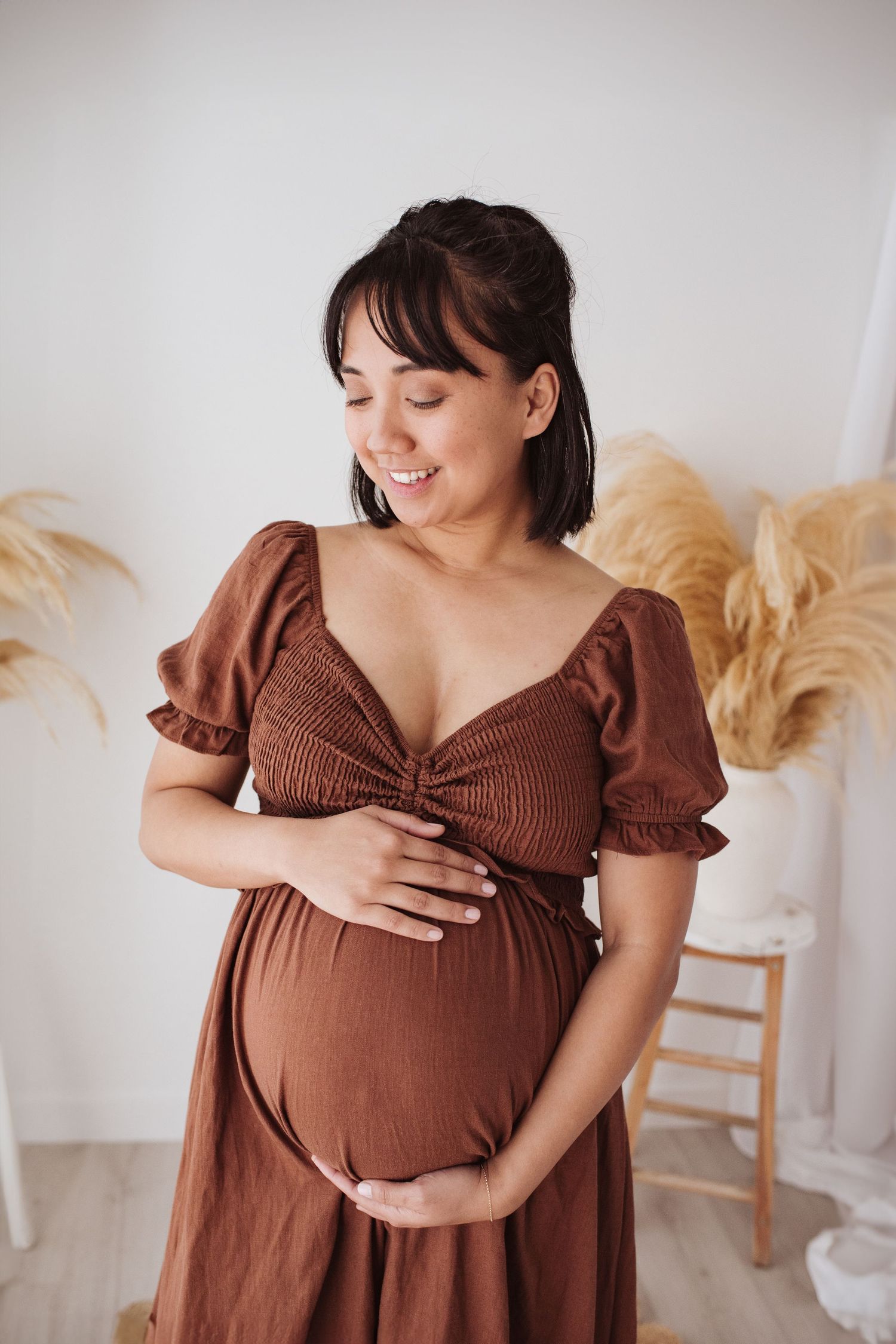 Expectant mother in brown puff sleeve dress smiles while cradling baby bump in front of pampas grass decor.