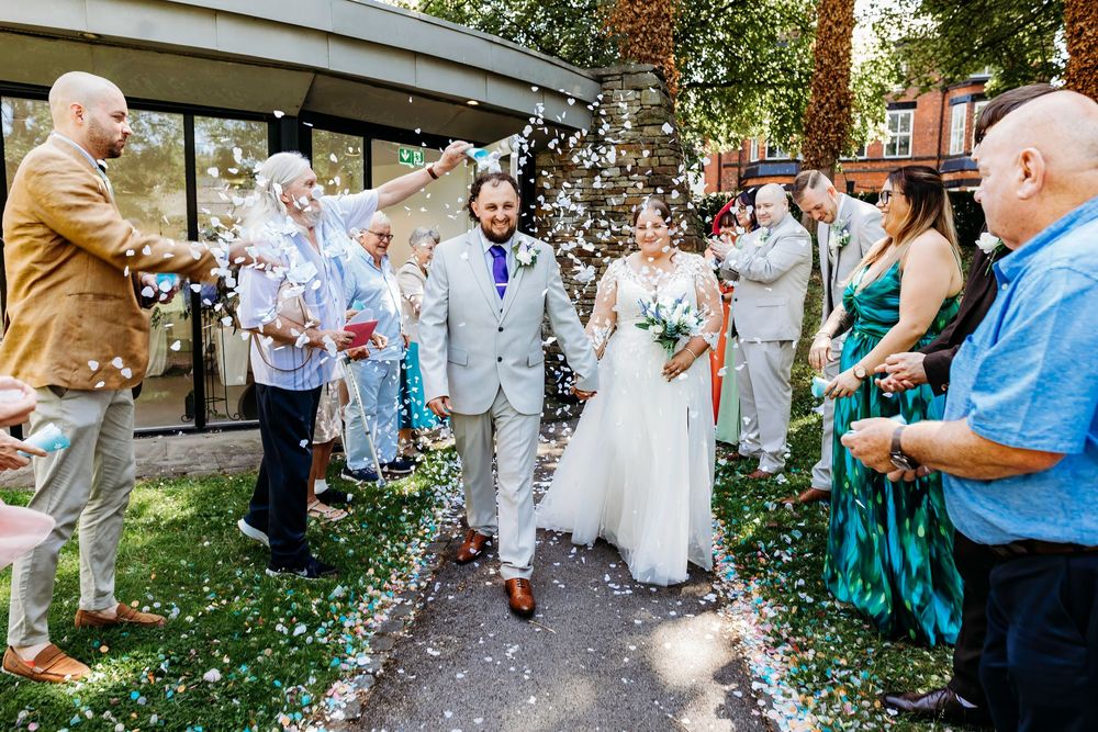 Wedding guests throw confetti as couple walks through garden path.