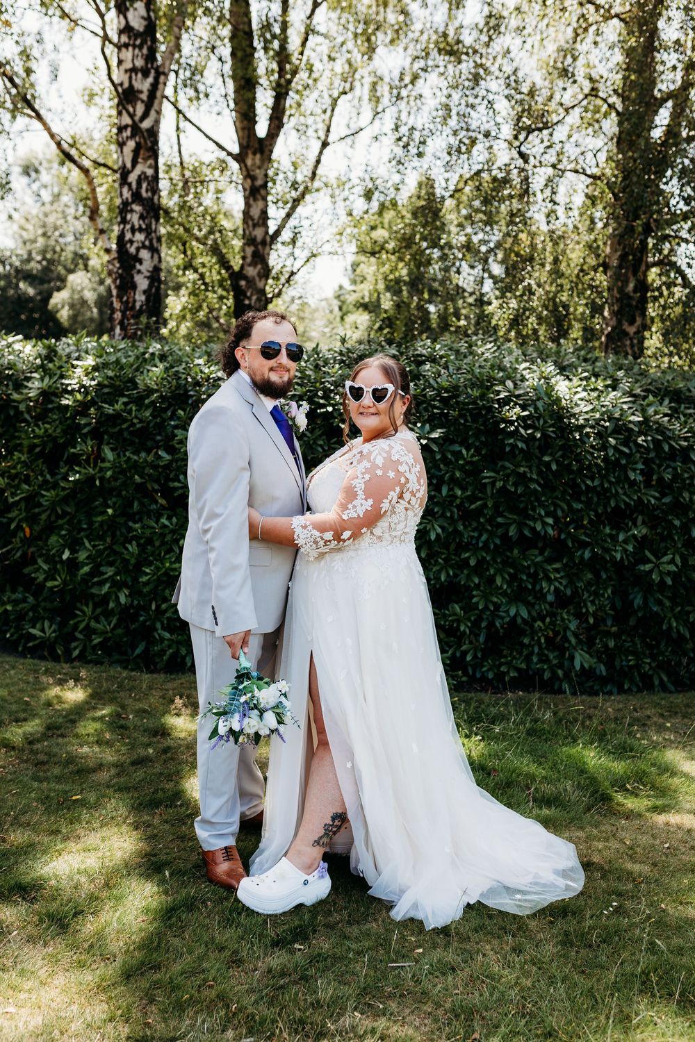 Child wearing white outfit sits beside person in formal wedding dress holding white bouquet in sunny garden.