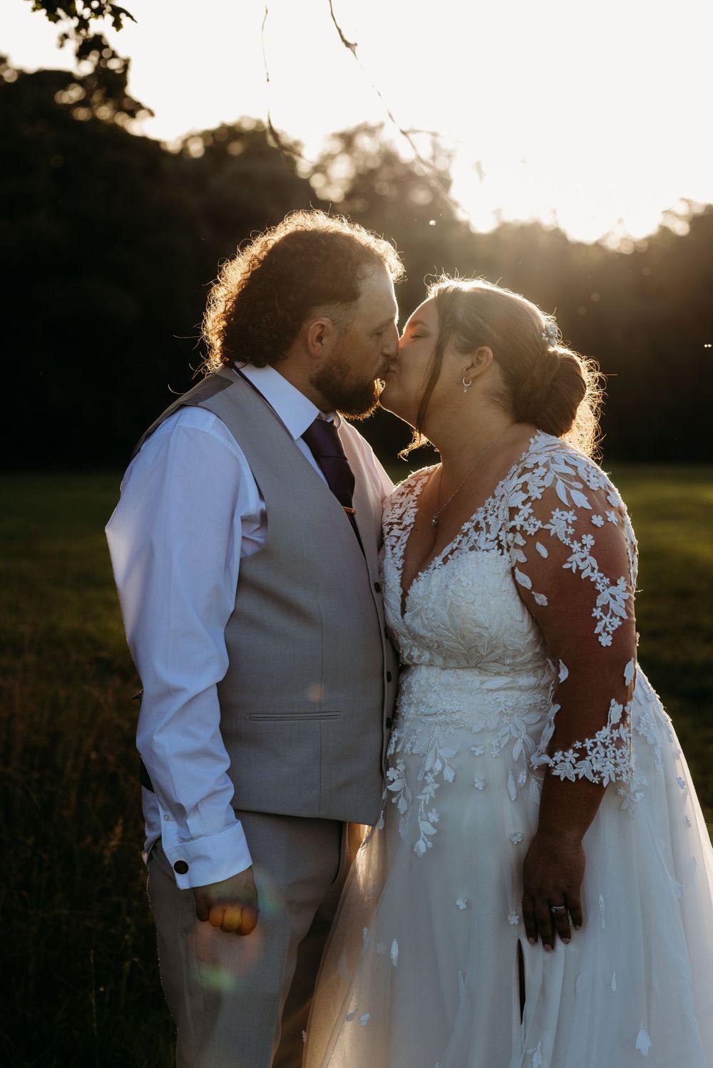 Newly married couple shares intimate sunset moment in garden setting with golden backlight and delicate lace dress details.