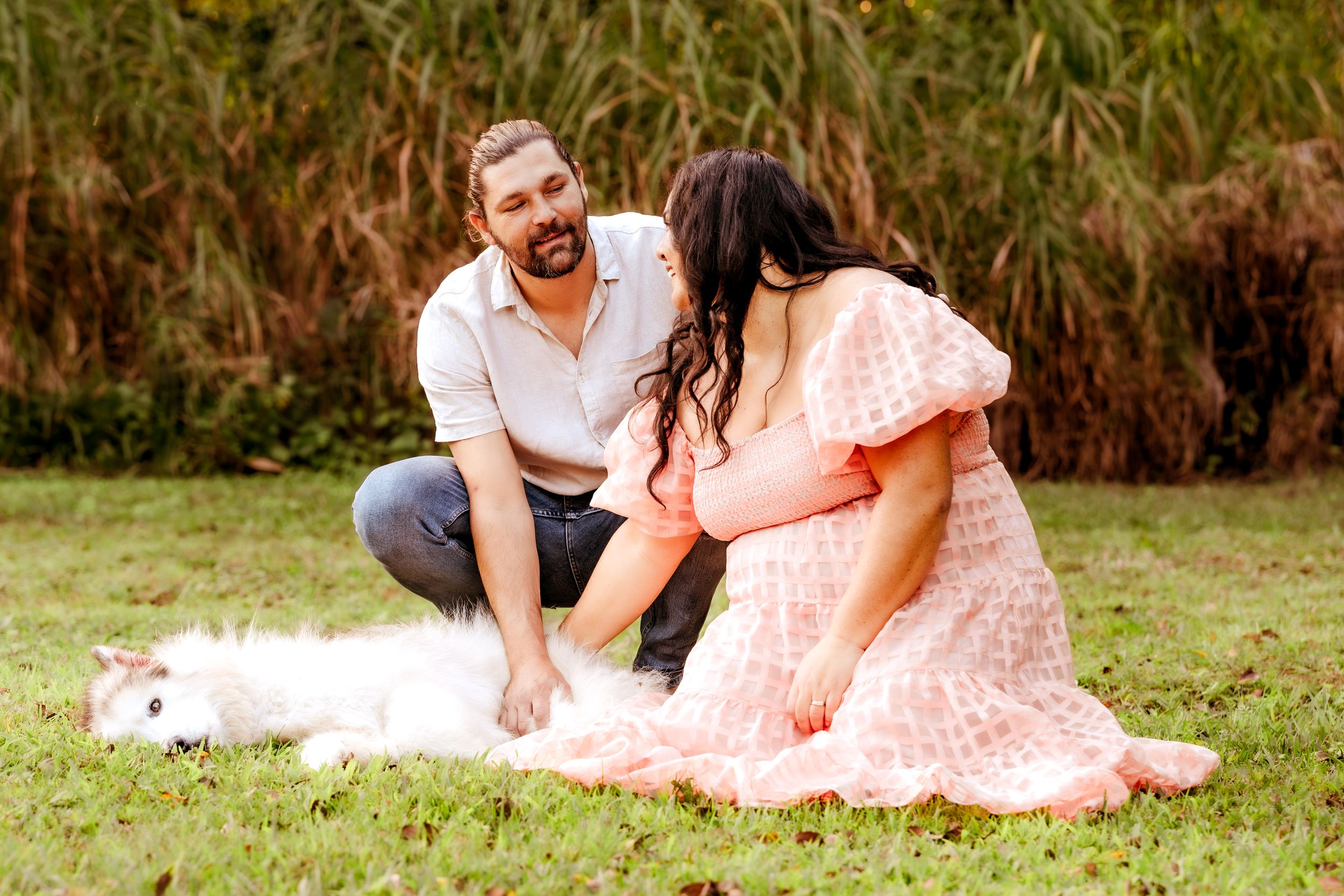 Couple in pink dress and white shirt playing with a fluffy white cat on green grass outdoors.
