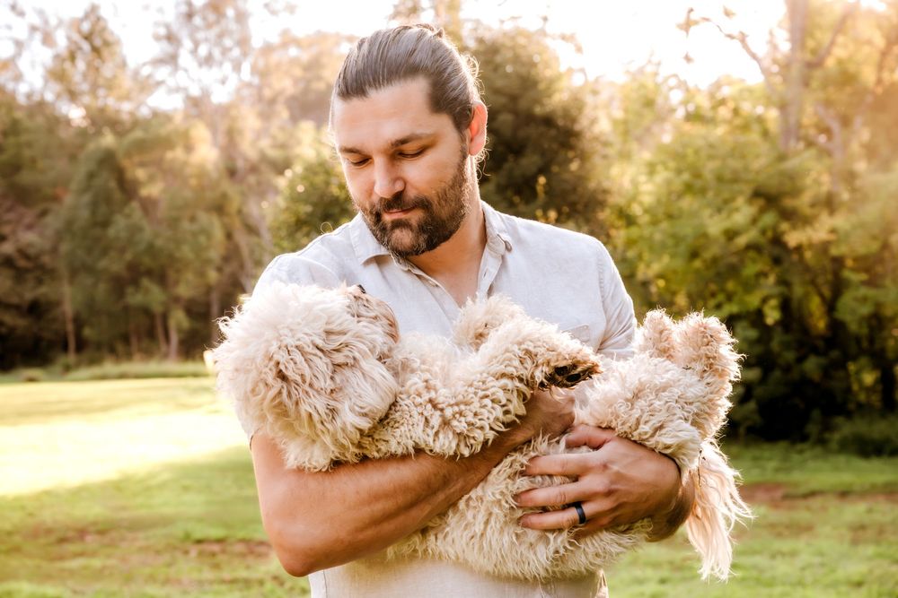 Bearded man with bun hairstyle holding a fluffy cream-colored dog outdoors in a lush green park setting.