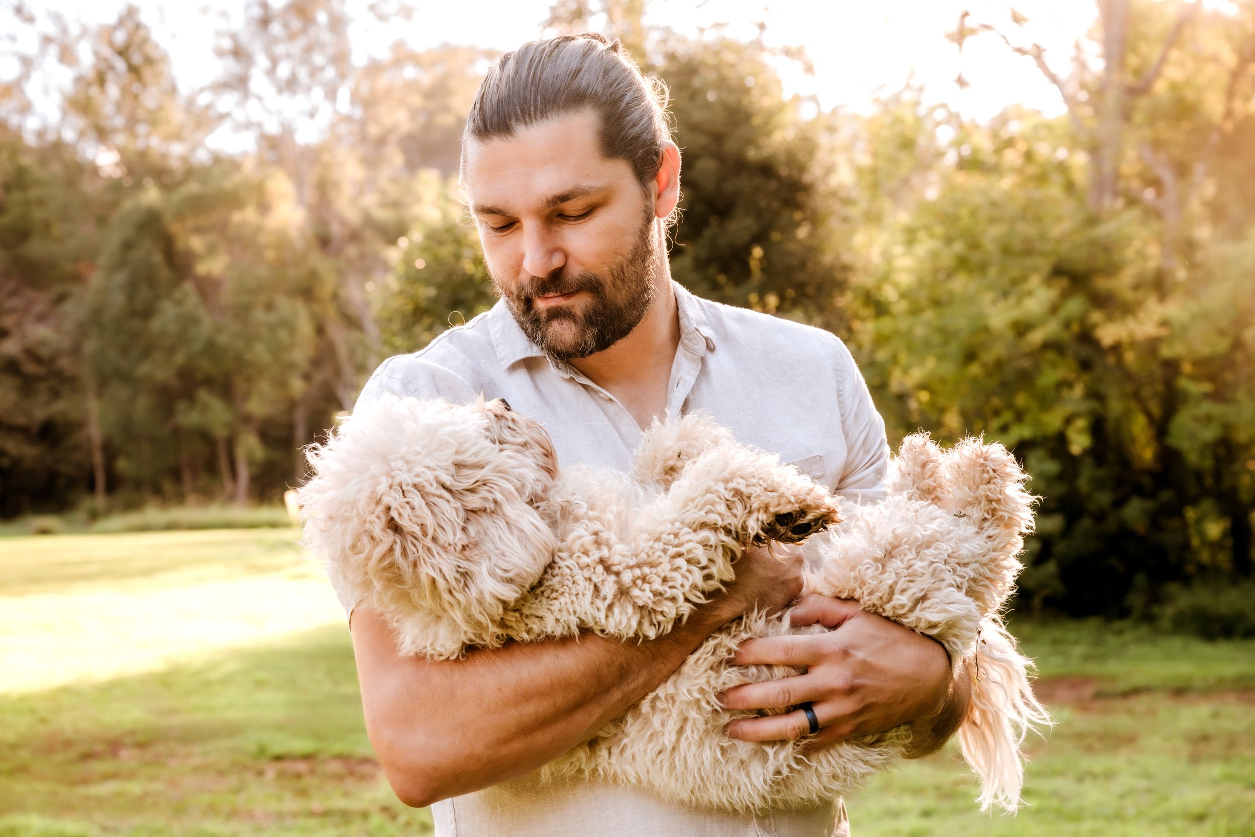 Bearded man with bun hairstyle holding a fluffy cream-colored dog outdoors in a lush green park setting.