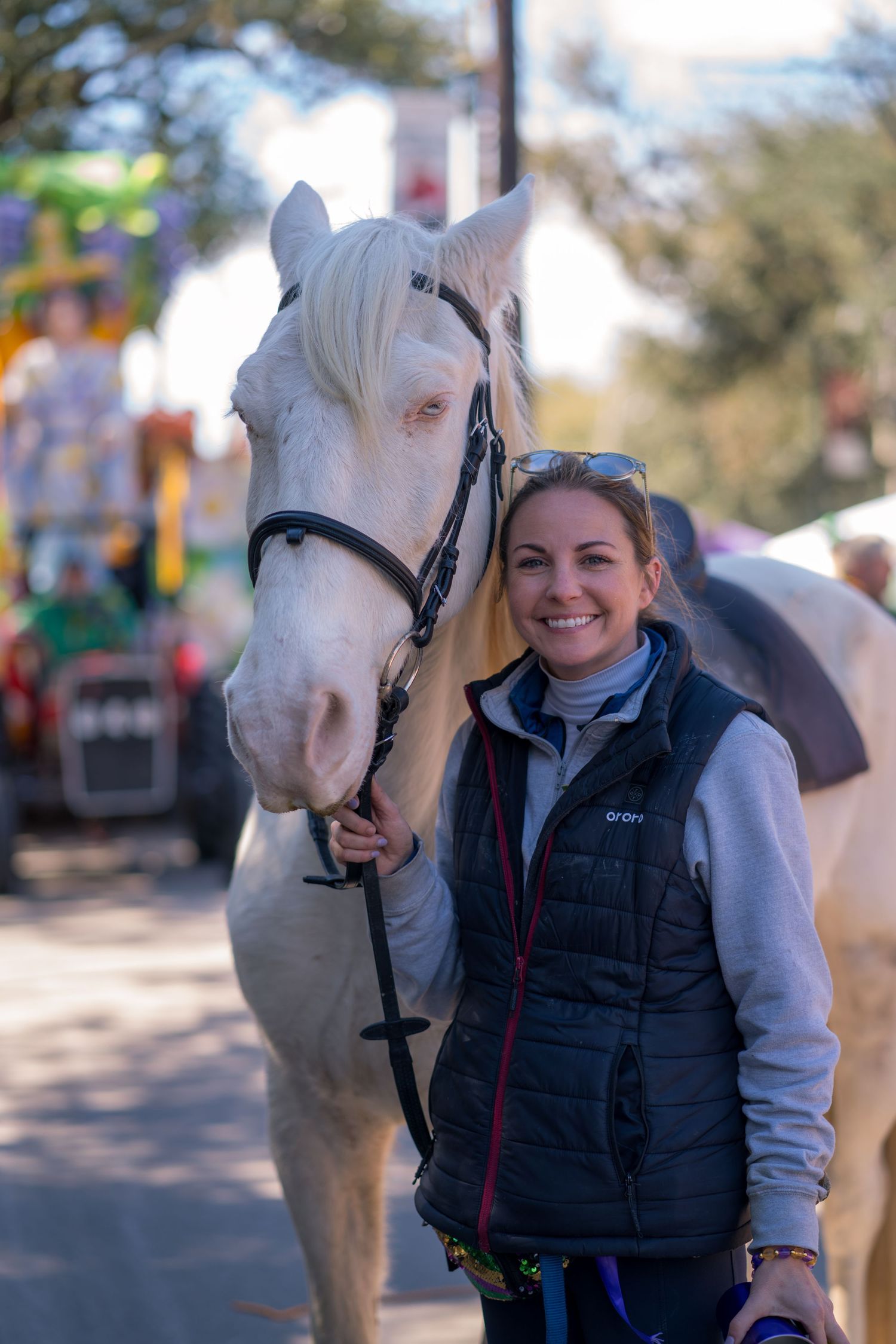 A white horse poses with someone in a black vest and gray sweater on a sunny day.