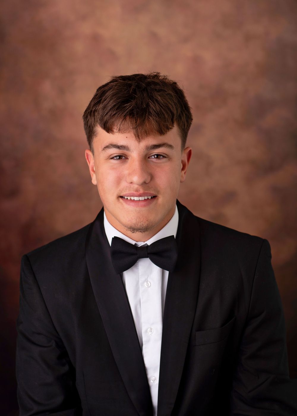 Young man in black tuxedo and bow tie poses for formal portrait photo against brown studio backdrop.