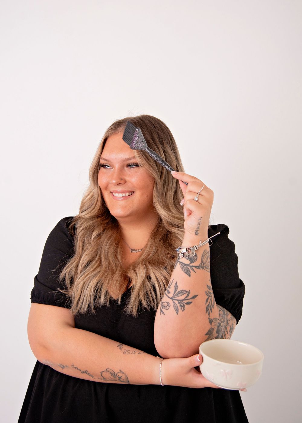 Woman in black dress holds hairbrush and smiles while styling blonde waves, showing arm tattoos against light background.