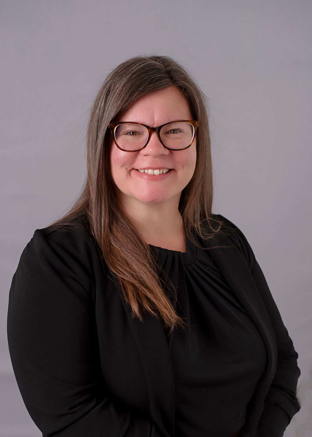 Professional headshot photograph of a smiling person wearing glasses and a black long sleeve top against a gray background.