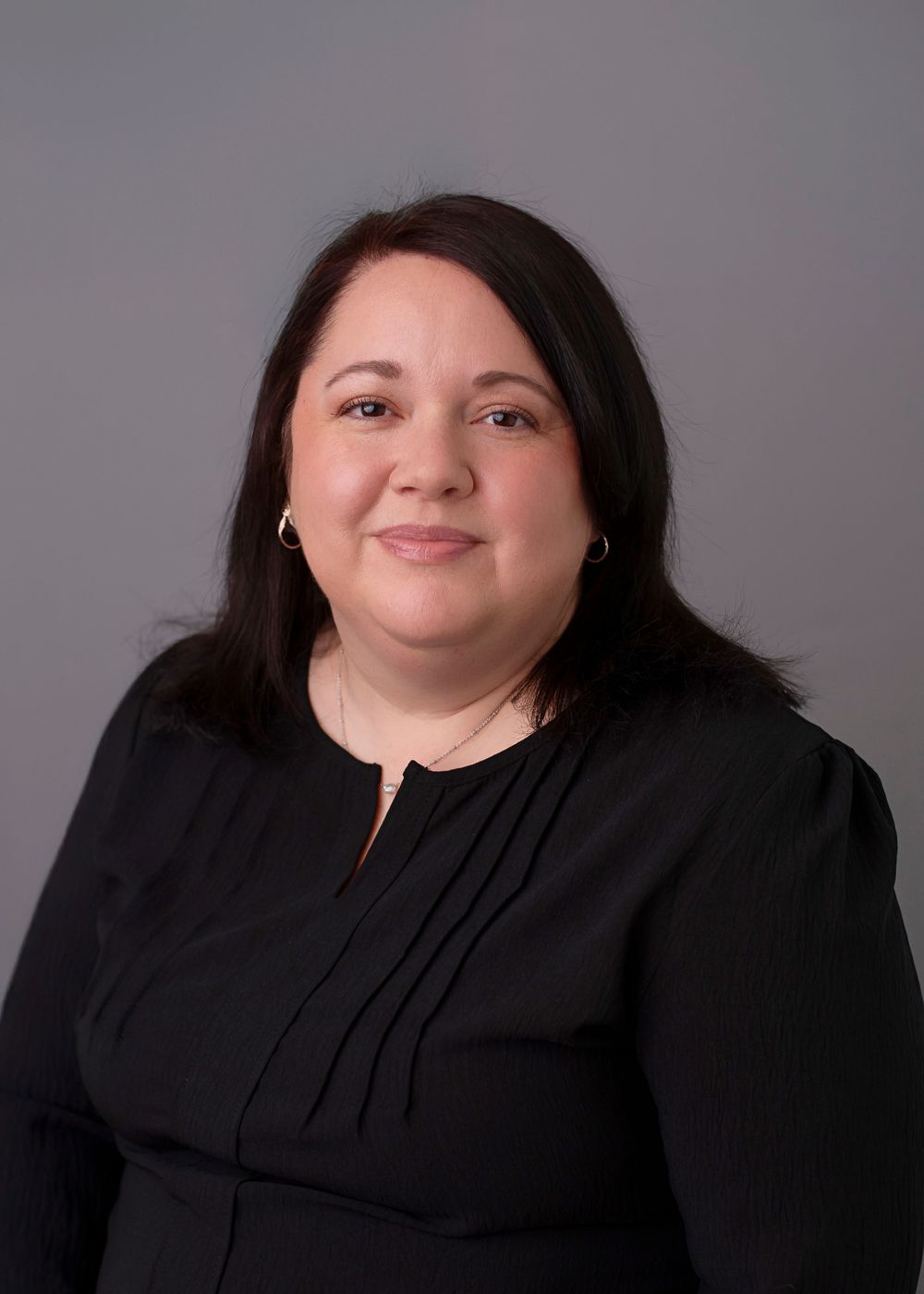 Professional headshot photo of someone in a black top with pleated neckline details against a gray background.