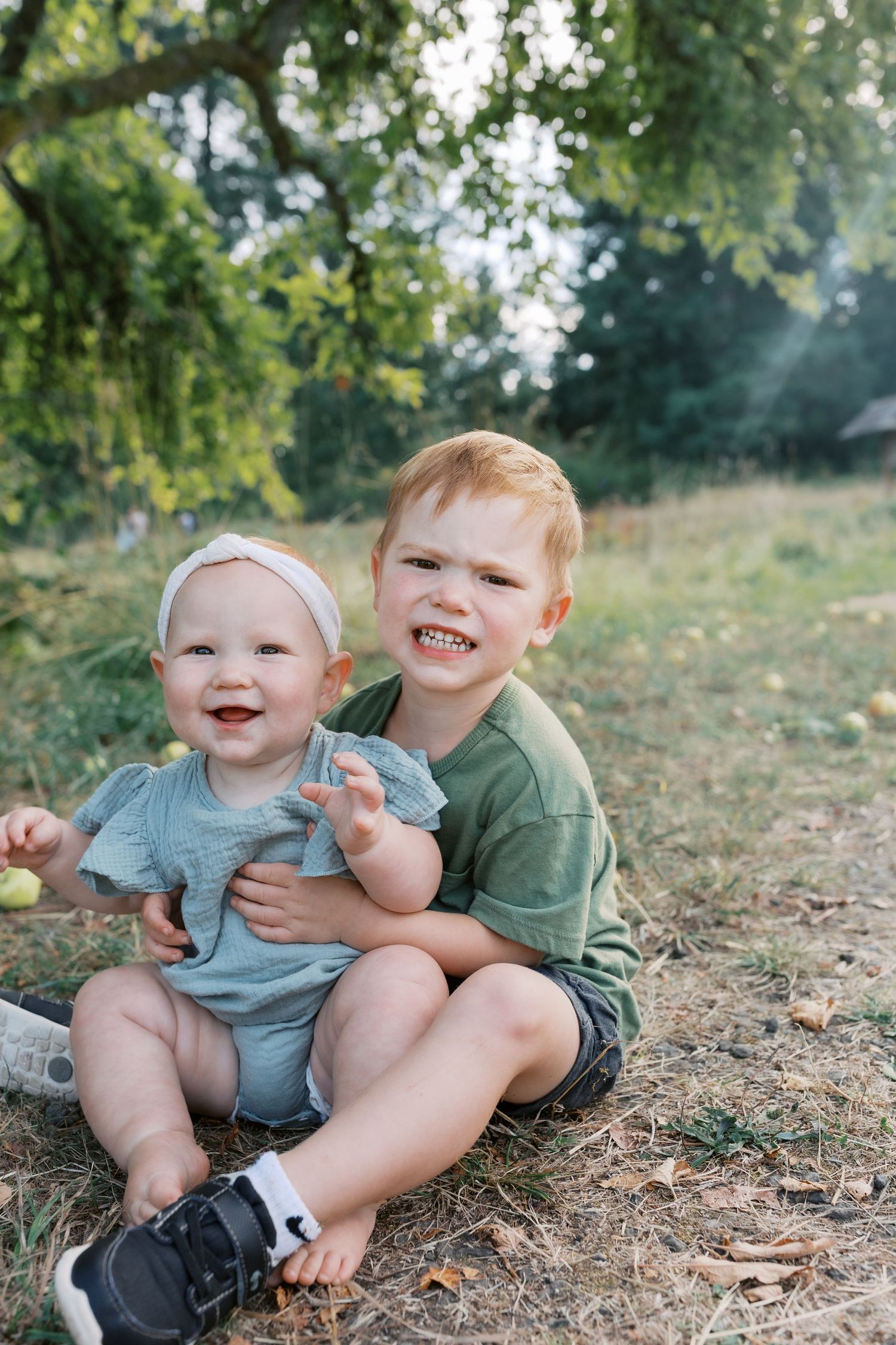 A smiling toddler girl and red-haired boy sitting together outdoors under a green tree on a sunny day.