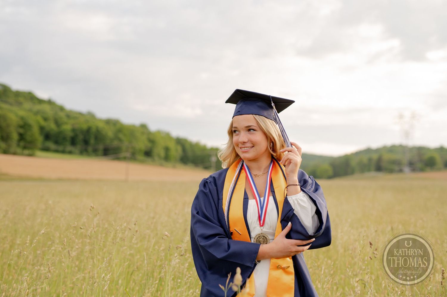 Graduate in navy cap and gown with medal and gold stole smiles against rural landscape.
