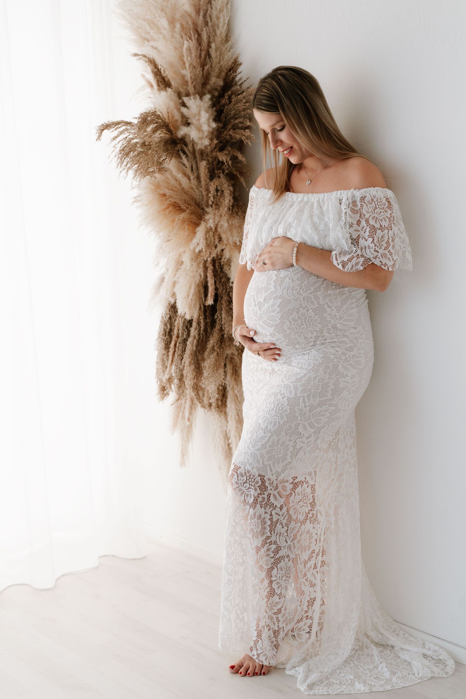 Pregnant woman in elegant white lace off-shoulder gown holding flowers beside bright window.