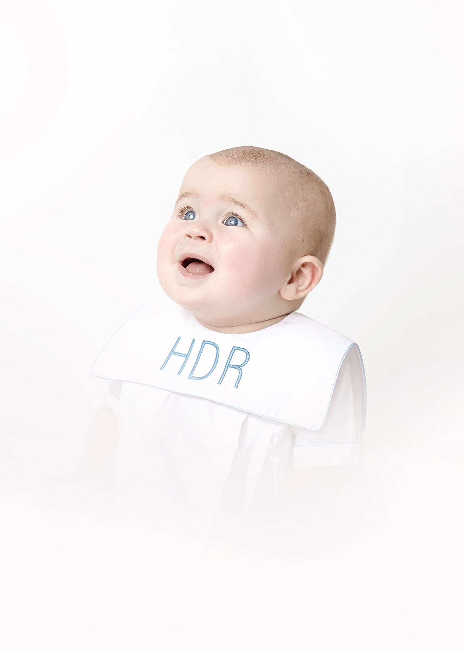 Baby in white collared outfit with HDR monogram looking upward against white background.