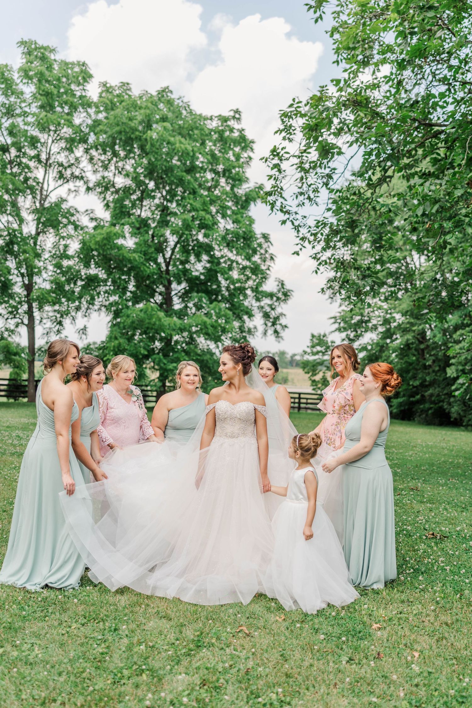 Bridesmaids in mint green dresses gather around bride on lush green lawn.