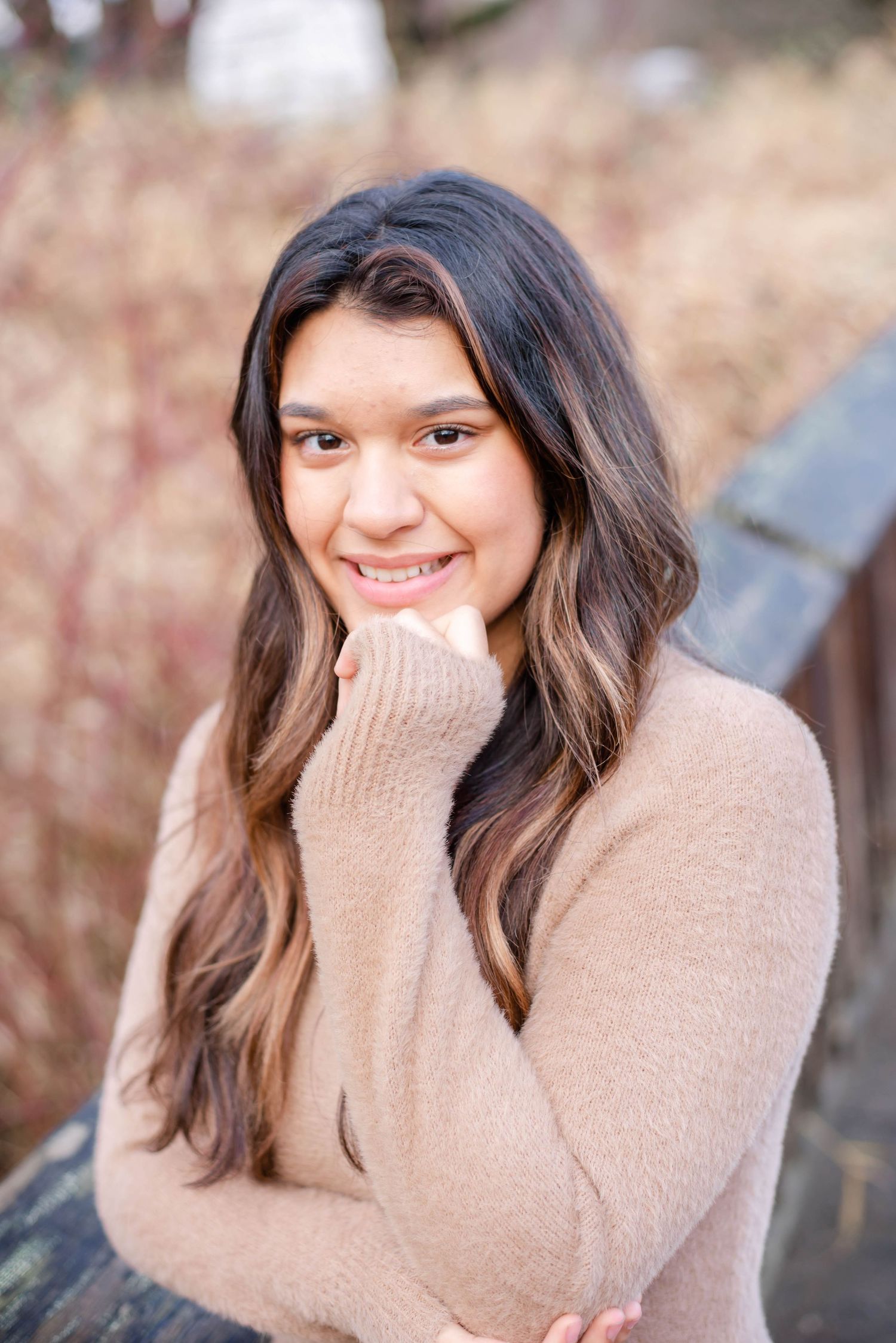 Person in beige sweater poses with hand near face against natural outdoor setting.