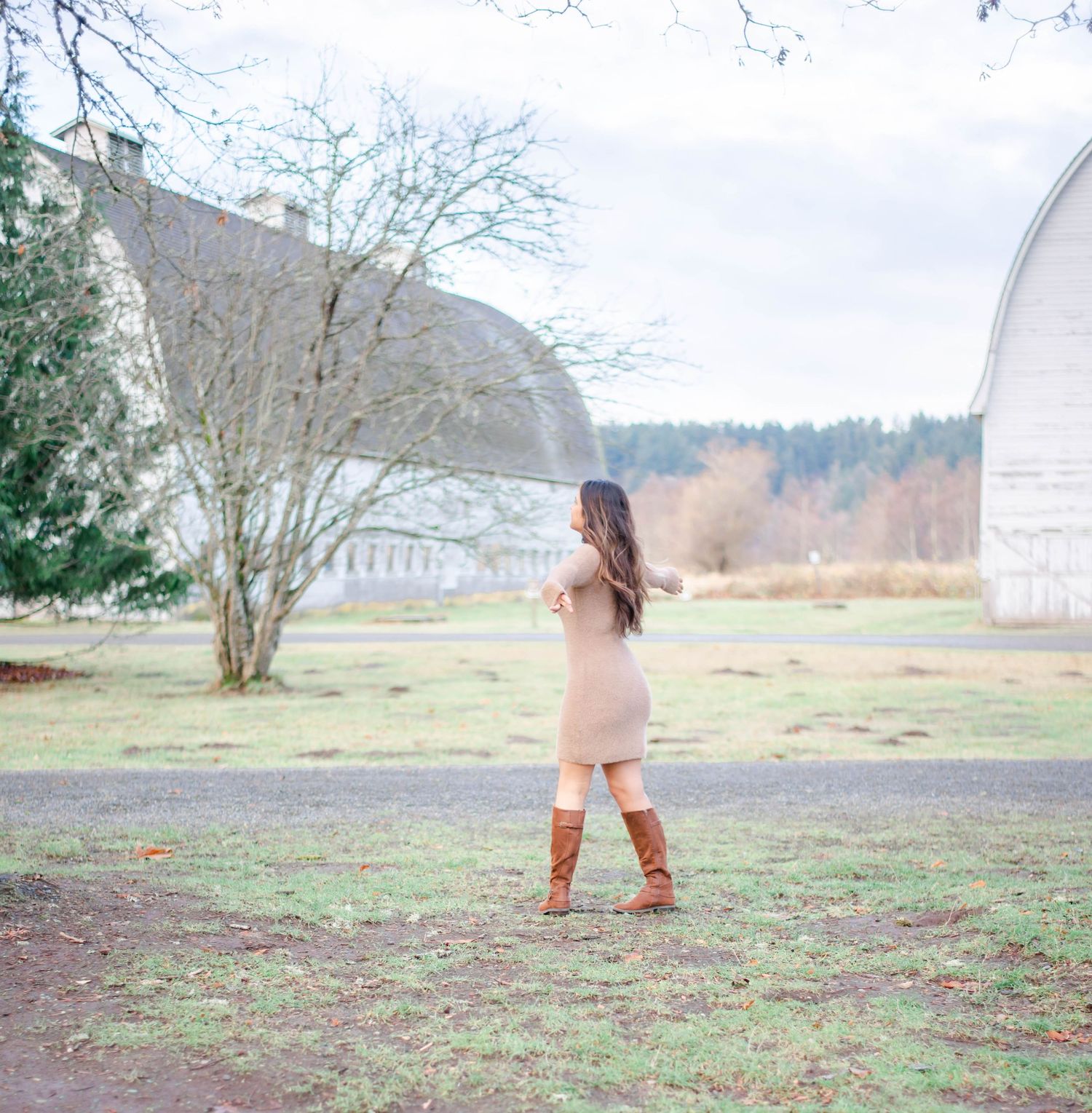 Figure in beige outfit stands in open field with bare trees and buildings in background.